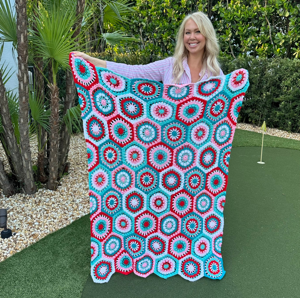 Picture taken outside of a blonde woman holding up a handmade crochet blanket. The blanket is crocheted using pink, red, turquoise, teal and white yarn. There are several crochet blocks in this blanket shaped as hexagons with one of each color in each block.